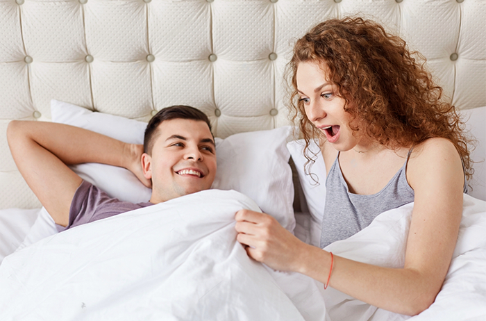 Shocked female looks under white blanket, being surprised with size of boyfriend`s penis after taking an Erectimus instant erection pill