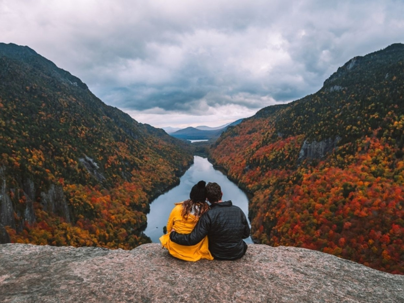 Two people sitting on a rock overlooking a scenic view of a lake surrounded by mountains with autumn foliage.